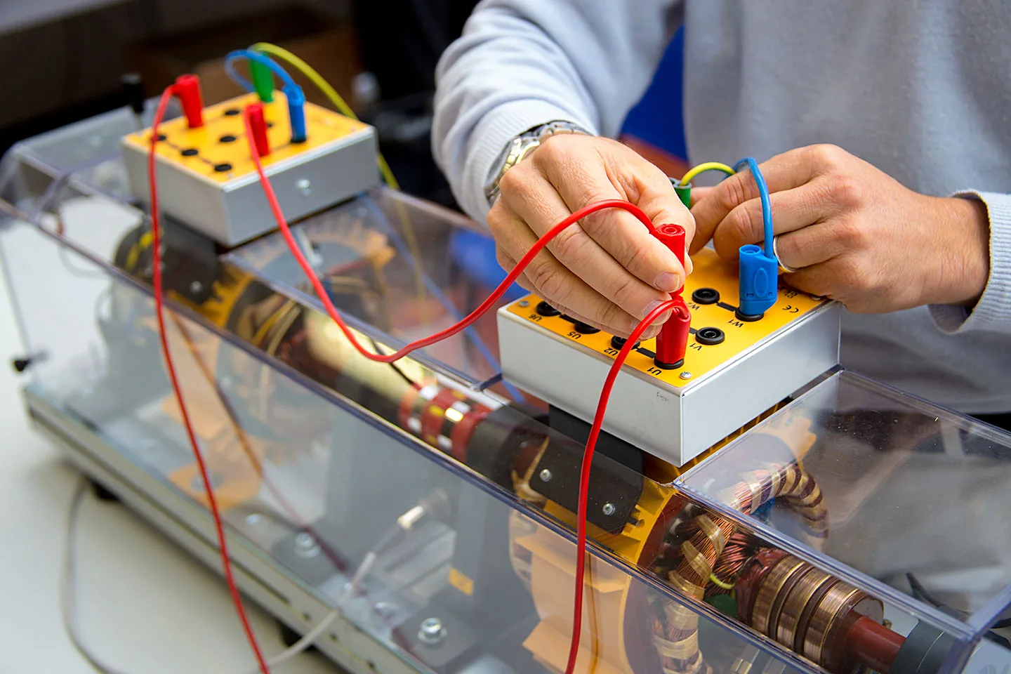 Manos conectando cables a la caja de bornes de una máquina eléctrica didáctica transparente