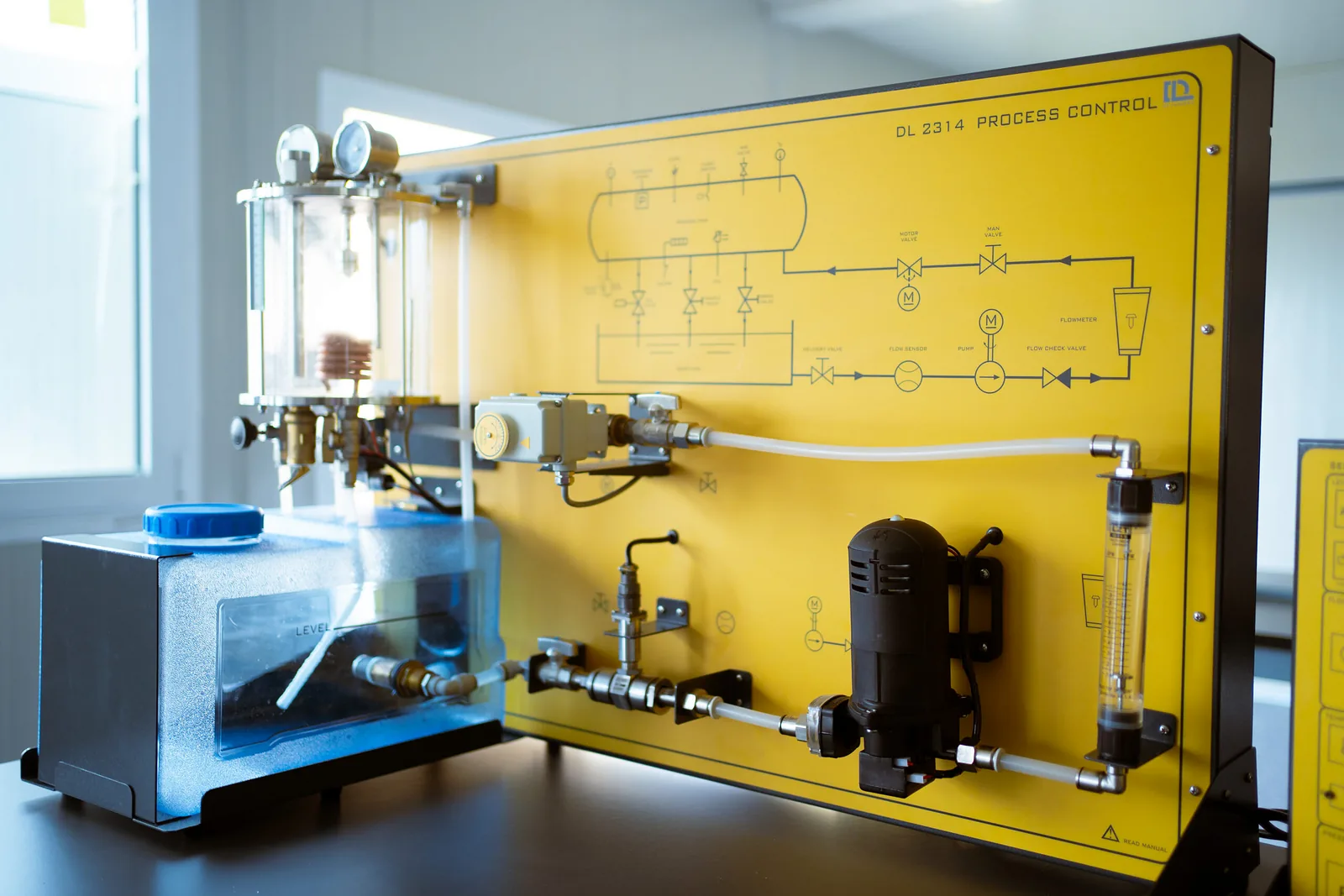 Entrenador didáctico de control de procesos con tanques transparentes, bomba e instrumentos en un panel amarillo